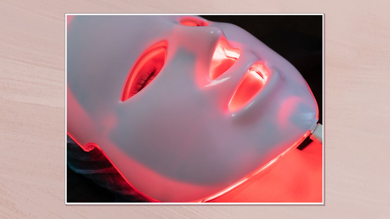 Image of a close up of a woman wearing an LED mask, in a white frame against a dusky pink watercolour-style background