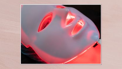 Image of a close up of a woman wearing an LED mask, in a white frame against a dusky pink watercolour-style background