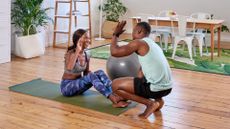 woman and man in an open plan home setting with wooden floors. they're on a green exercise mat, wearing gym clothes about to give each other a high five.