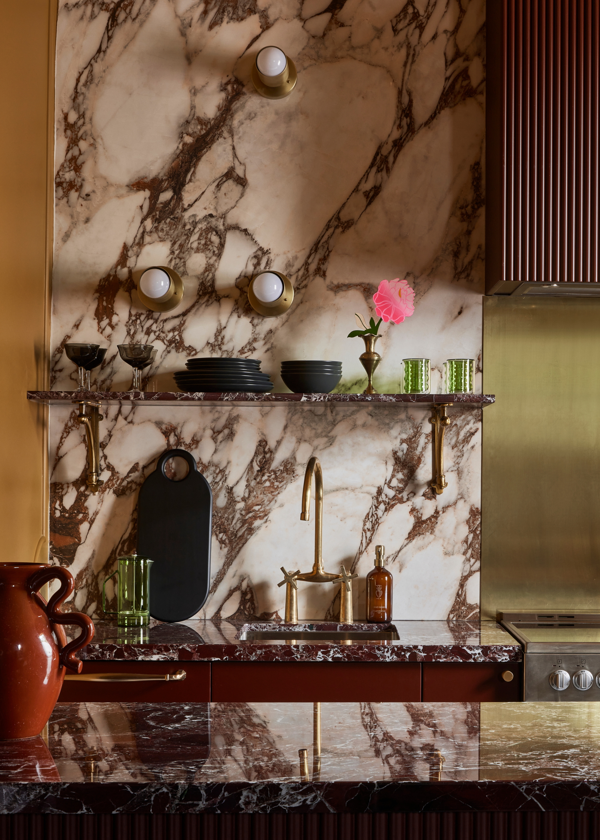 A mixed material backsplash in a burgundy marble kitchen with two red and white marbles, brushed brass, a fluted extractor hood and three small brass wall lights