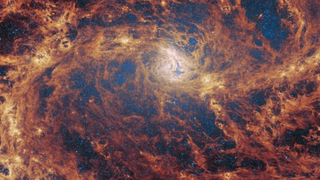 A close-up view of a barred spiral galaxy. Two spiral arms reach horizontally away from the core in the centre, merging into a broad network of gas and dust which fills the image. This material glows brightest orange along the path of the arms, and is darker red across the rest of the galaxy. Through many gaps in the dust, countless tiny stars can be seen, most densely around the core.
