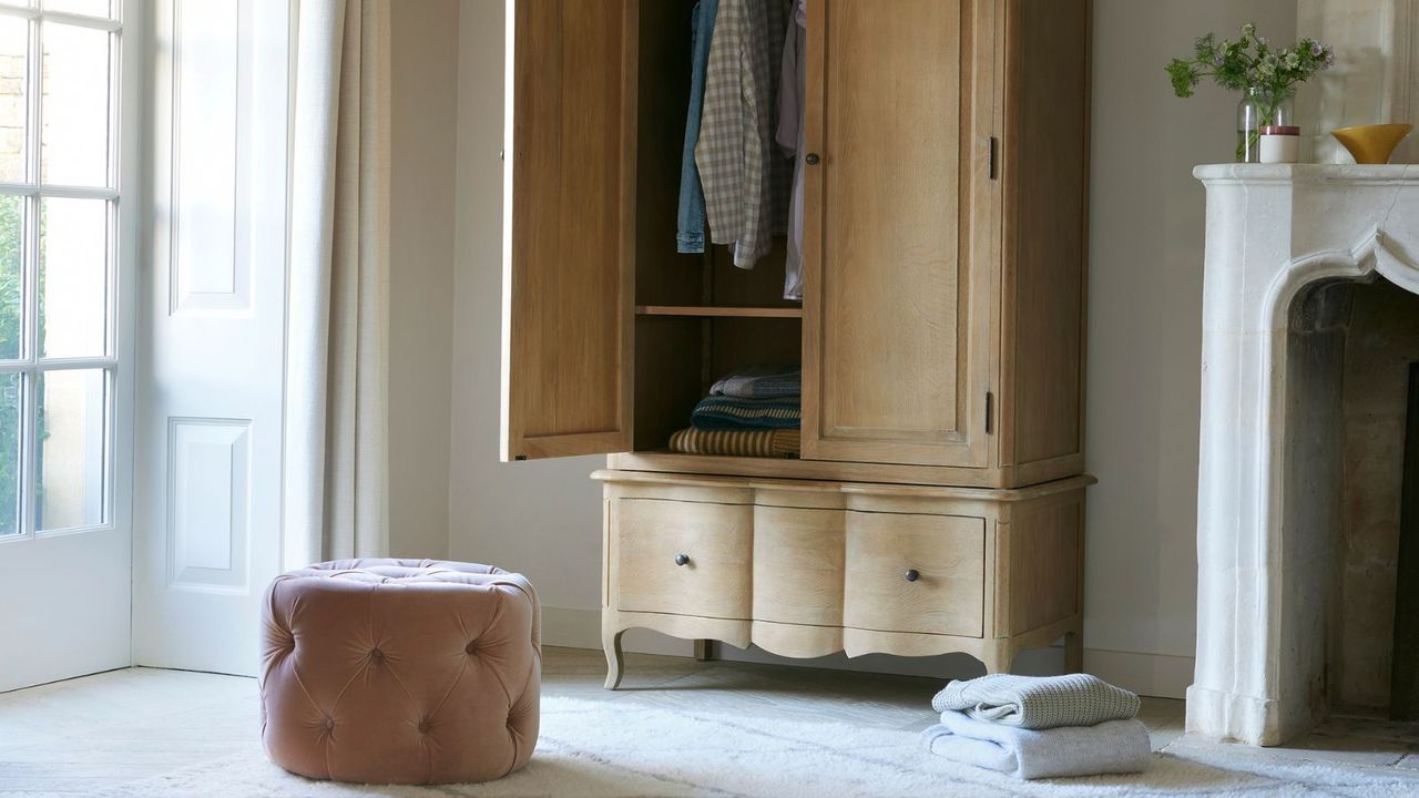 Loaf closet half open with a pink pouffe seat in front of it and a small pile of folded clothes on the floor beside it. 