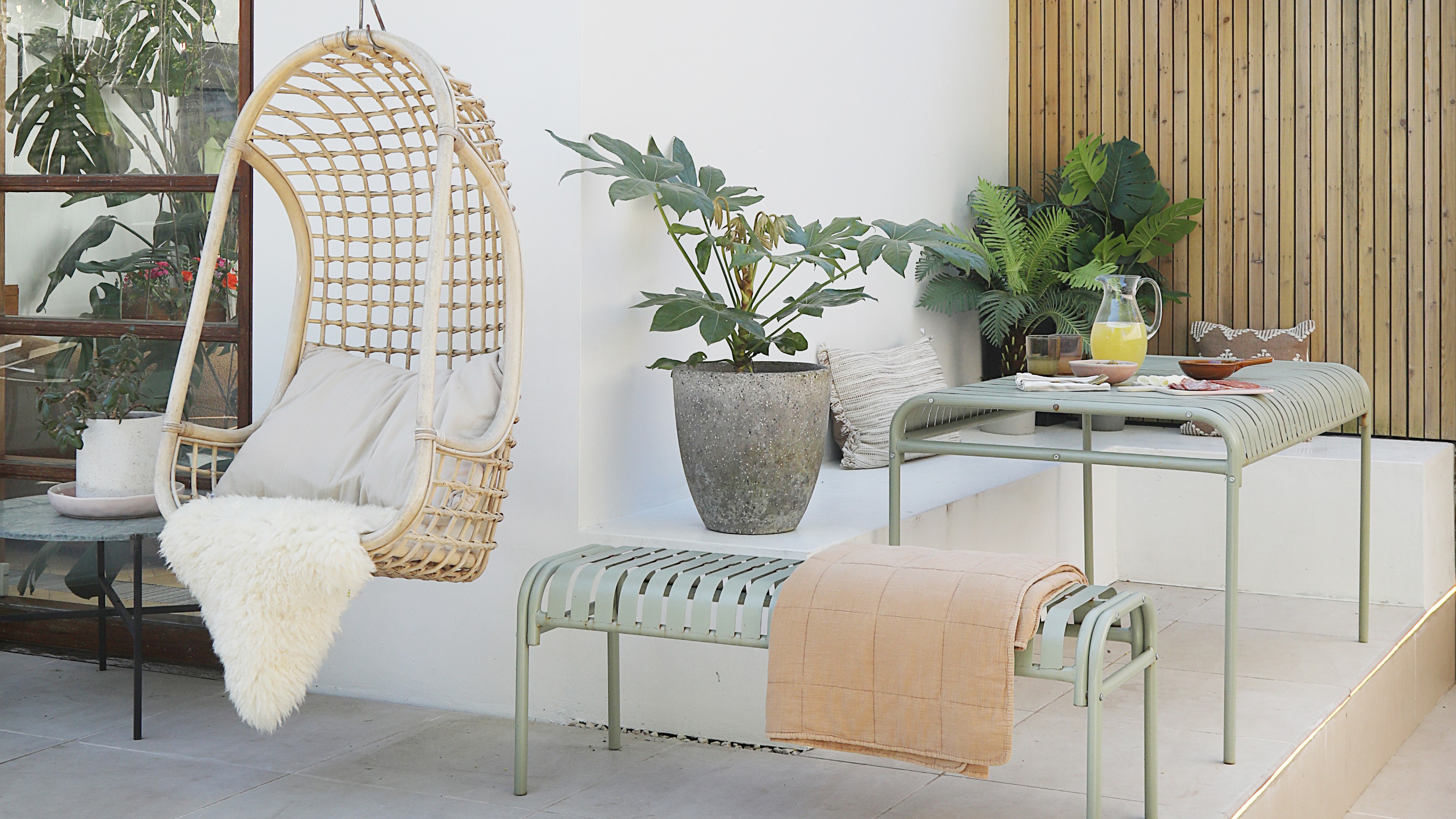 A patio with a sage green HAY Palissade metal outdoor dining set with ahanging rattan egg chair next to it
