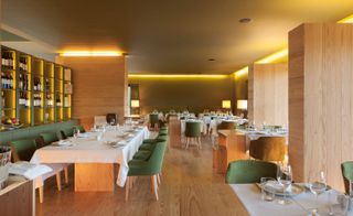 The dining area in the Hotel Monverde. The Interior is made up of light wood. To the left, we have a muted green shelf with wine on it. Tables are arranged throughout the room, made of the same light wood, with white linen and velvet green chairs.