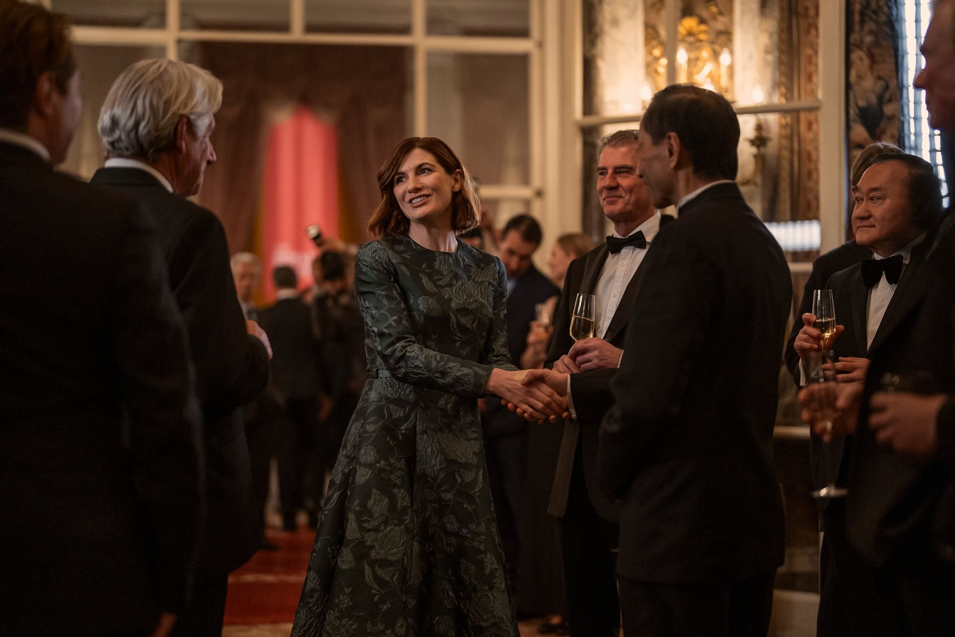 A woman in a green gown (Jodie Whittaker as Bethan Vaughn) greets men wearing tuxedos in a grand ballroom, in 'The Diplomat' season 4.