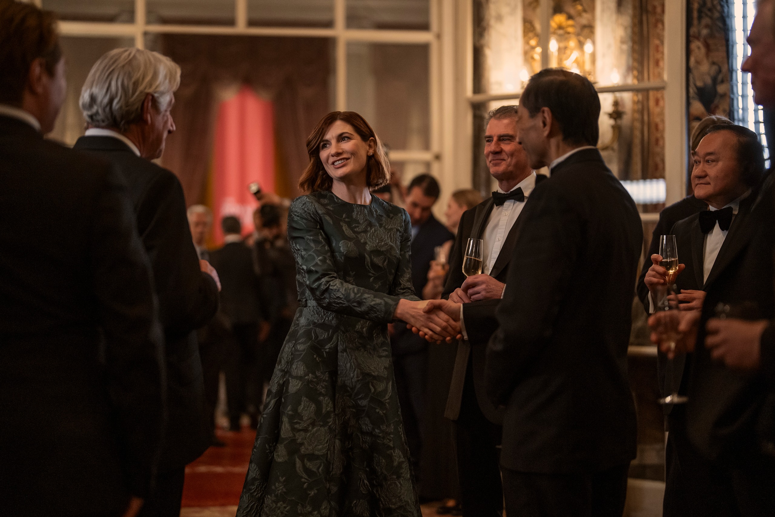 A woman in a green gown (Jodie Whittaker as Bethan Vaughn) greets men wearing tuxedos in a grand ballroom, in 'The Diplomat' season 4.