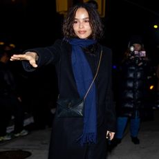 Zoe Kravitz wears a blue scarf, long black coat, ysl bag, black sheer tights, and black open-toe sandals.