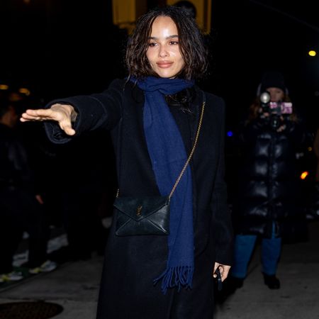 Zoe Kravitz wears a blue scarf, long black coat, ysl bag, black sheer tights, and black open-toe sandals.