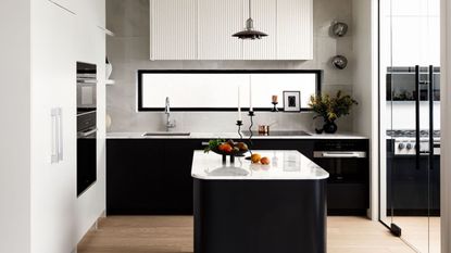 black kitchen with white stone countertops, gray backsplash, oak timber floors, white cabinetry, fluted range hood, window above stove, and glass doors leading to working kitchen