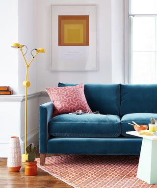 A living area with white walls with wall art and windows, a deep blue velvet couch with a pink throw pillow, a tall yellow metal lamp, a wooden floor with a peach rug and white geometric table on it