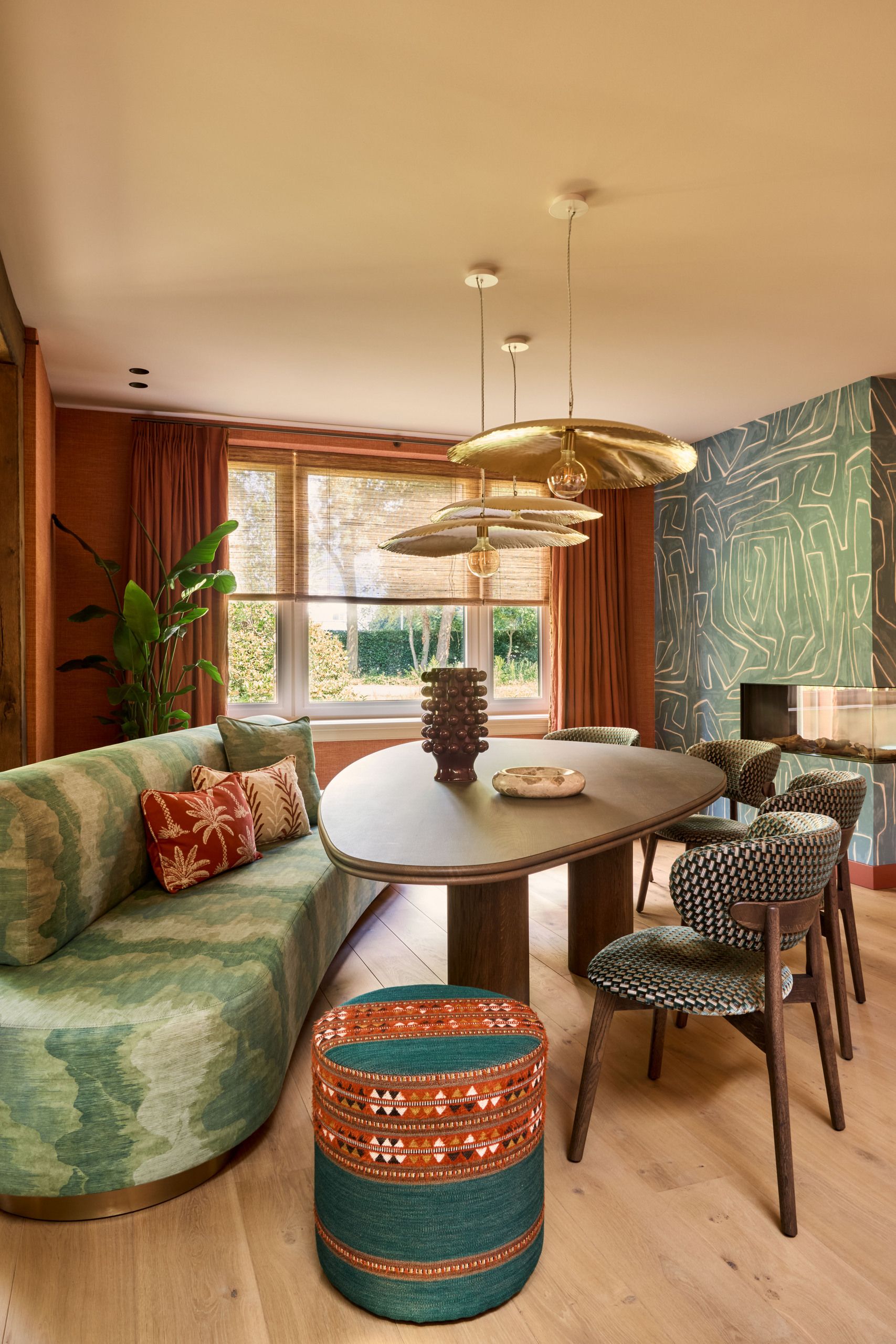 Image of a modern and colorful dining area with a teal patterned bench seat, an organically-shaped table, four chairs, a teal and orange pouffe, and orange and teal walls.