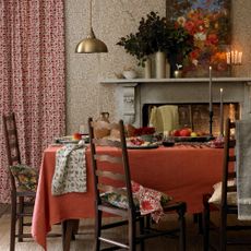 An autumnal dining room in candle light with a burnt orange tablecloth on the table and botanical beige wallpaper on the walls