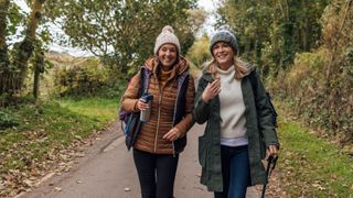 Two mature women wearing warm, casual, outdoor clothing and accessories walking along a tree-lined road as they talk and laugh together.