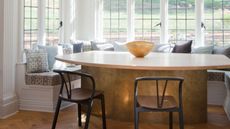 window seat dining area modern brass table with upholstered seat and cushions, and two wishbone chairs, parquetry floors, and pendant light overhead