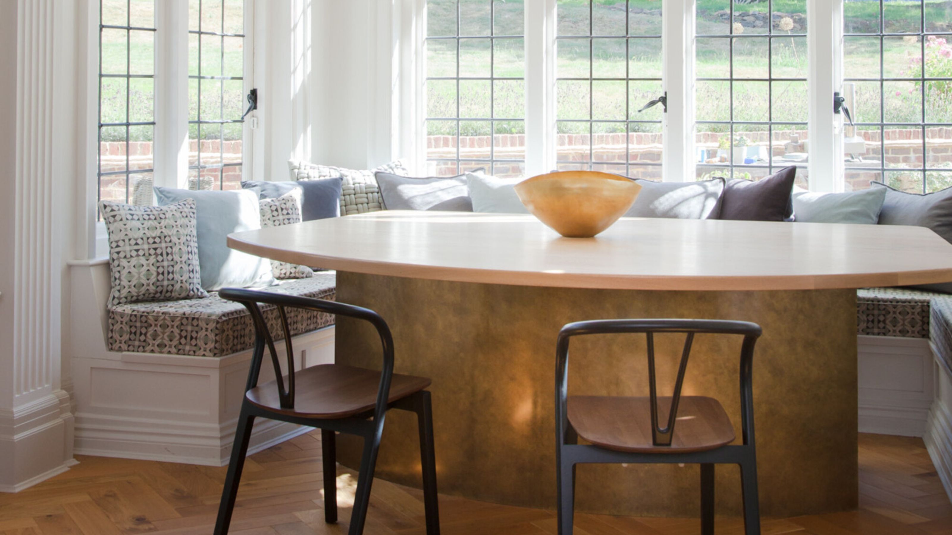 window seat dining area modern brass table with upholstered seat and cushions, and two wishbone chairs, parquetry floors, and pendant light overhead