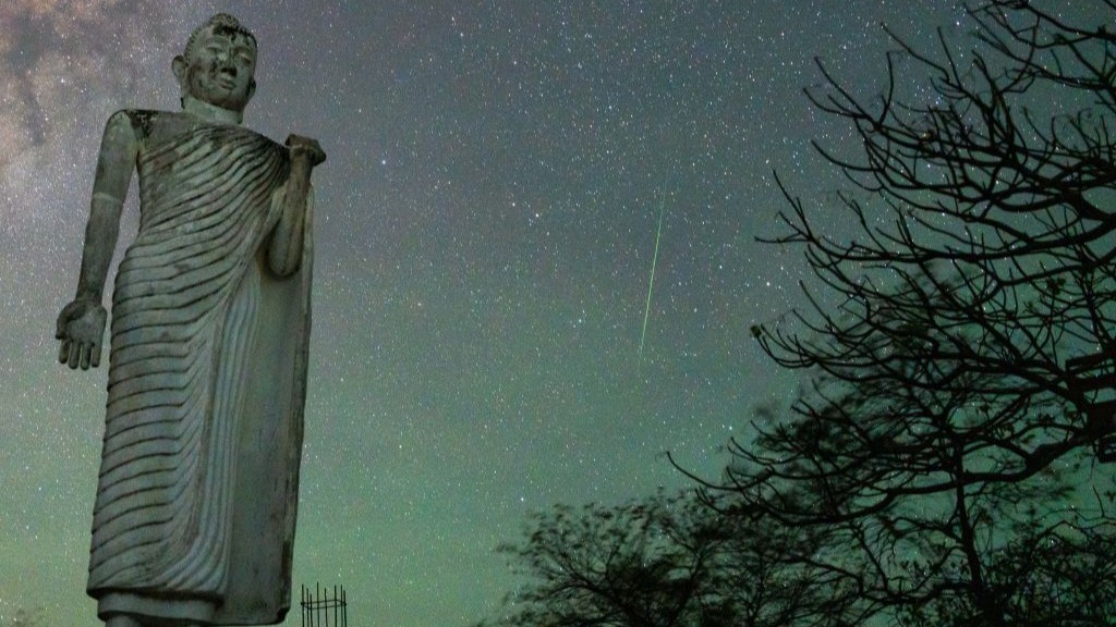 a large stone statue stands tall on the left , there is a tree on the right and a white streak of a meteor through the star filled sky between them.