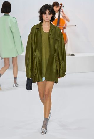 model wearing green coat over oversized shirt with gray socks and metallic shoes