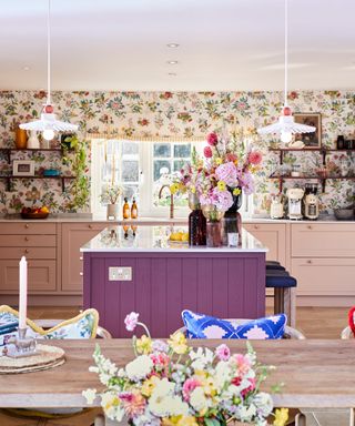 A colorful kitchen with floral wallpaper, pink cabinets, a purple kitchen island, two hanging pendant lights, and a table with three seats with colorful seat cushions