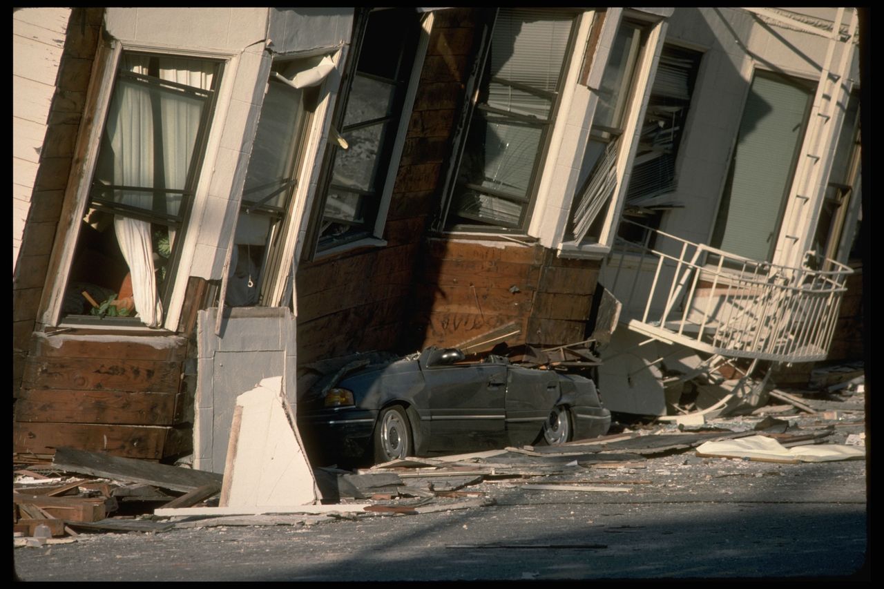 A gray car is partially visible under a tilted apartment building with a white curved balcony resting directly on the road behind it.