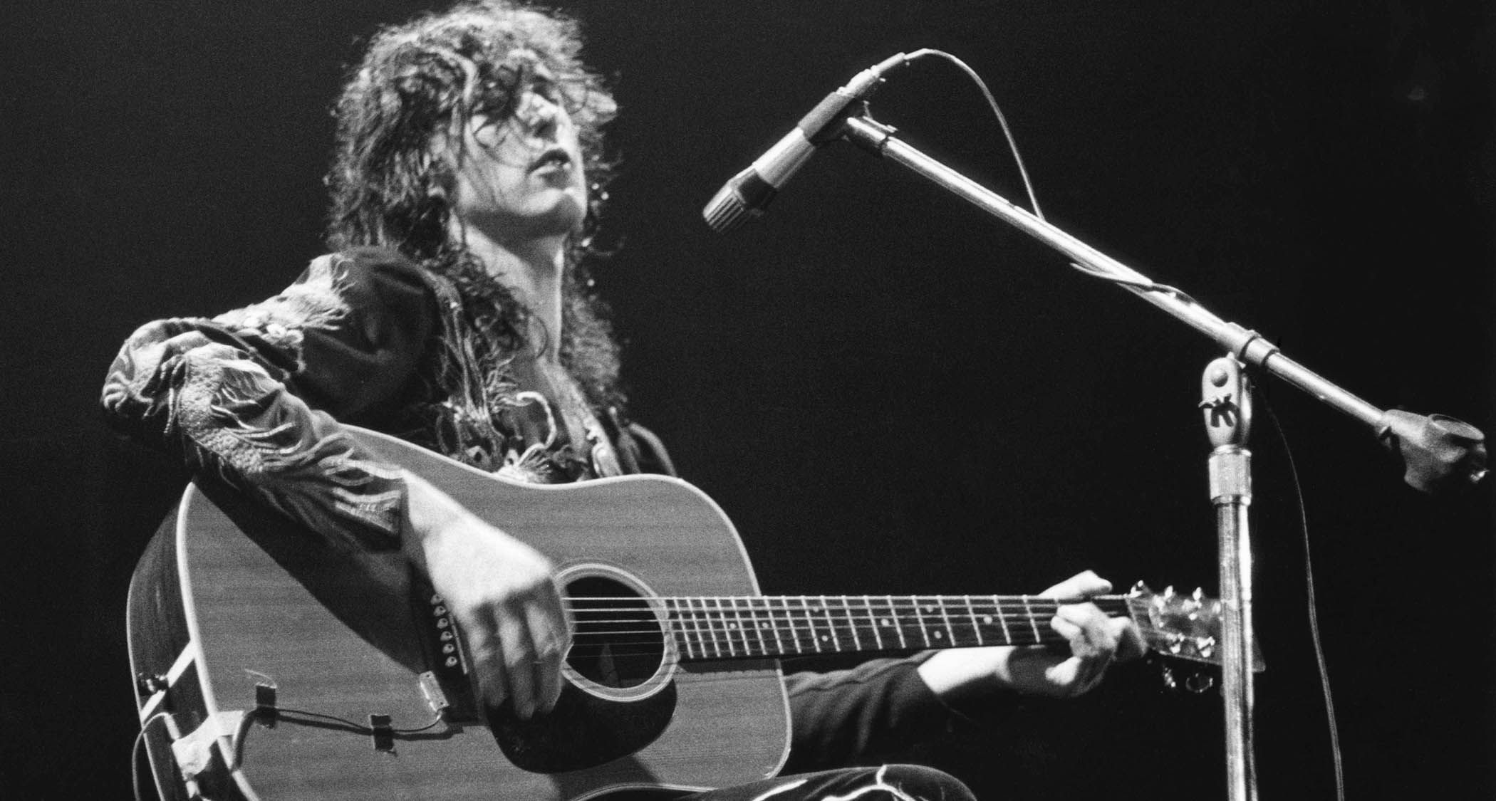 Jimmy Page plays a dreadnought acoustic, seated in front of a mic, in this B/W archive shot from Earl&#039;s Court, 1975.