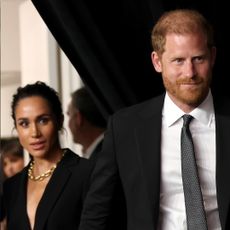 Meghan Markle wears a black dress and a large gold necklace and holds hands with Prince Harry, who has red hair and wears a black suit and white shirt