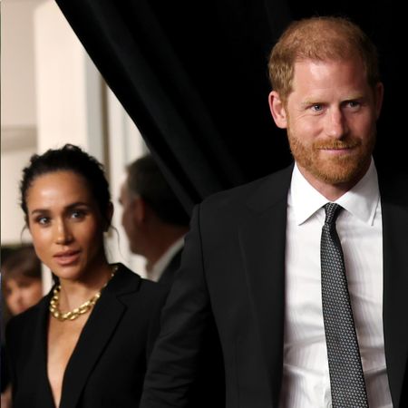 Meghan Markle wears a black dress and a large gold necklace and holds hands with Prince Harry, who has red hair and wears a black suit and white shirt