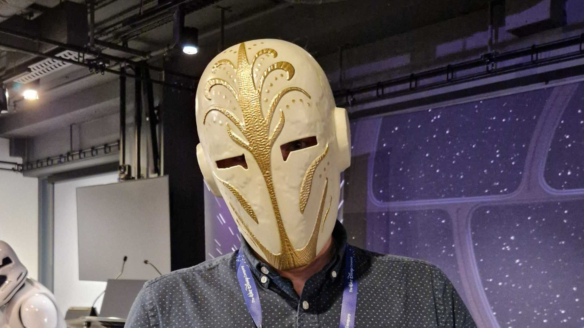 Someone wearing a Star Wars Jedi Temple Guard Mask against a star-filled background, with industrial features visible behind