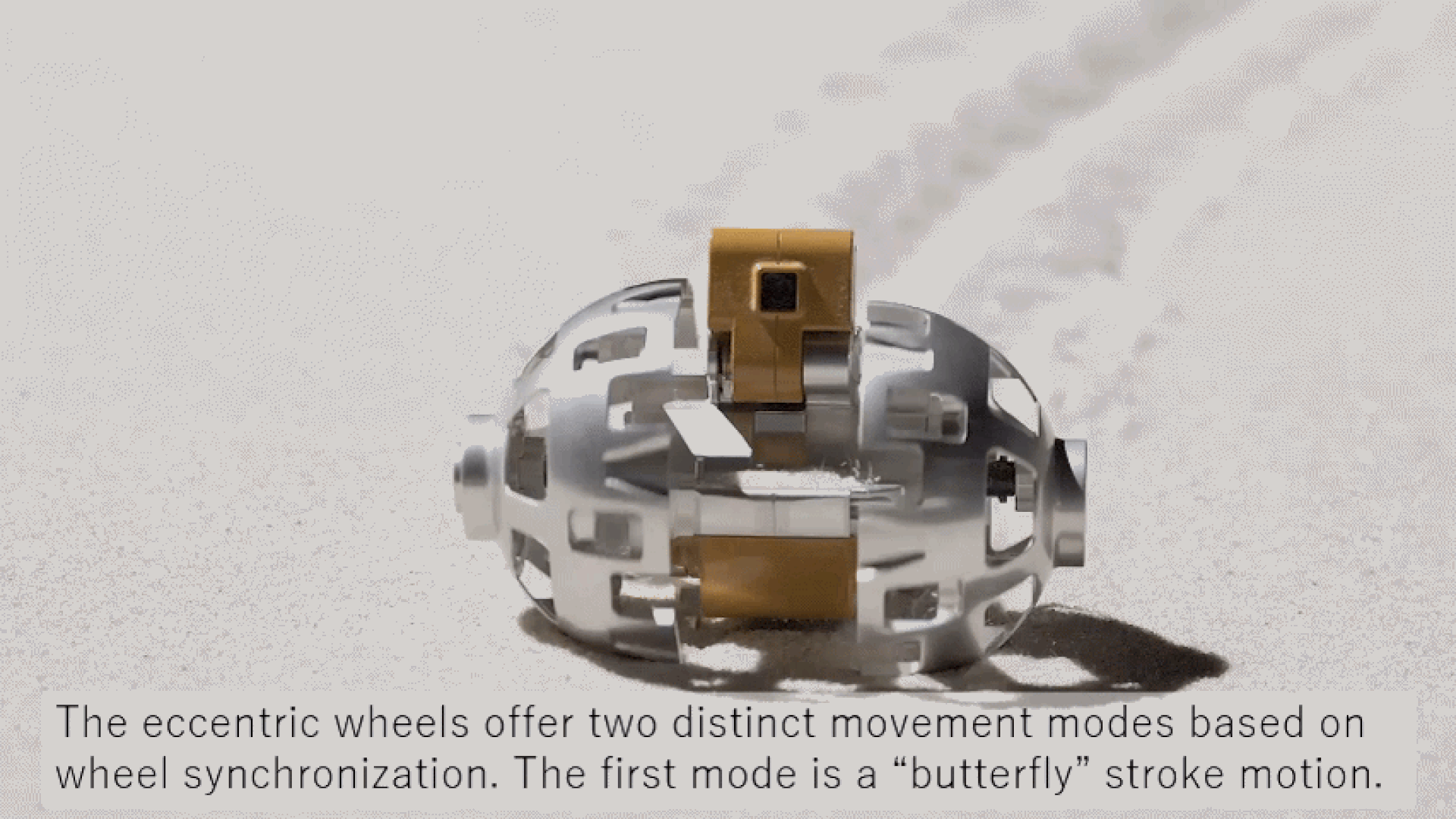 A close up of an oval shaped metal roller with a camera in the middle rolling on a sandy surface.