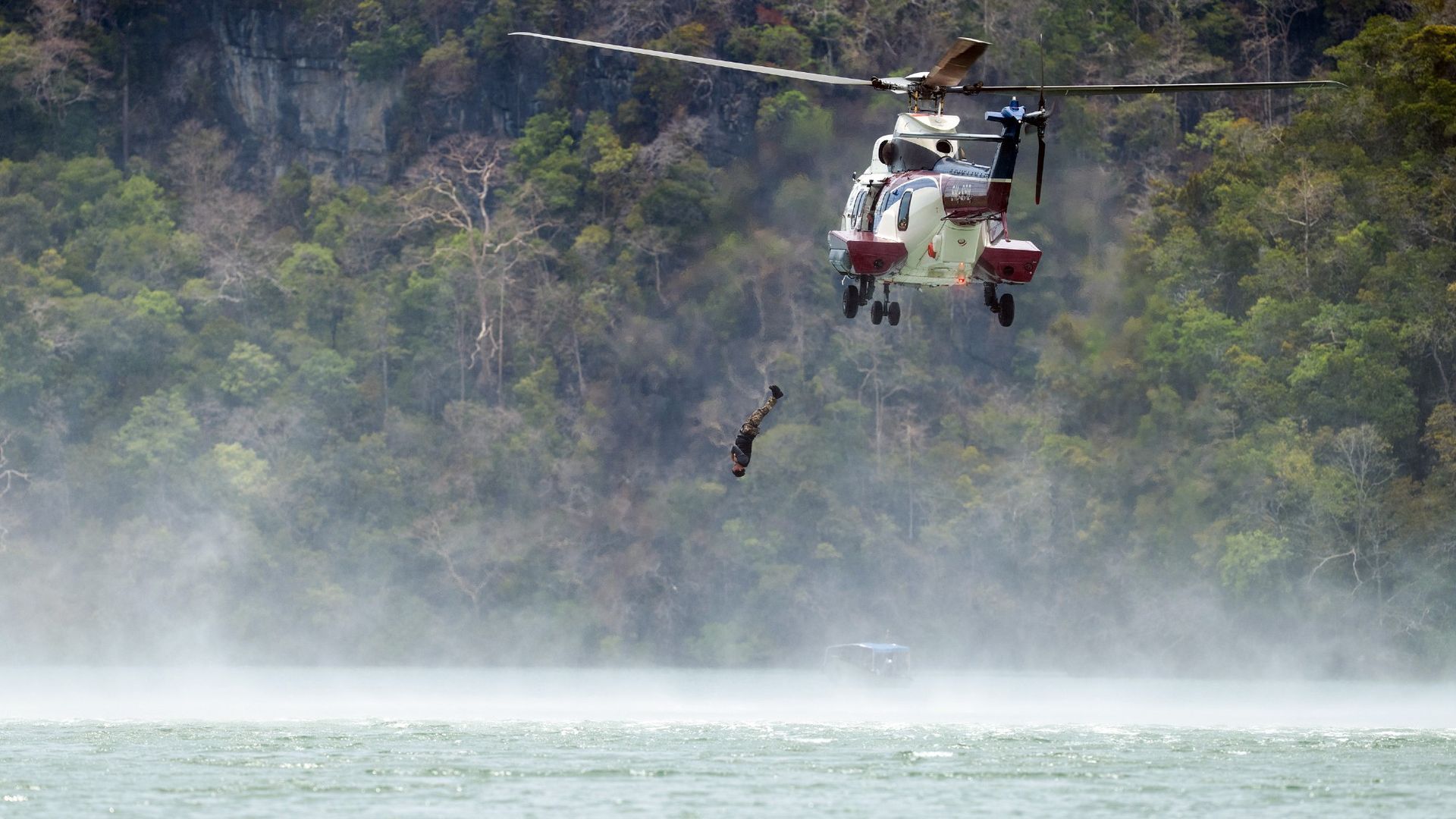 A recruit falls backwards out of a helicopter into the water. Trees are in the background and the water is light blue.