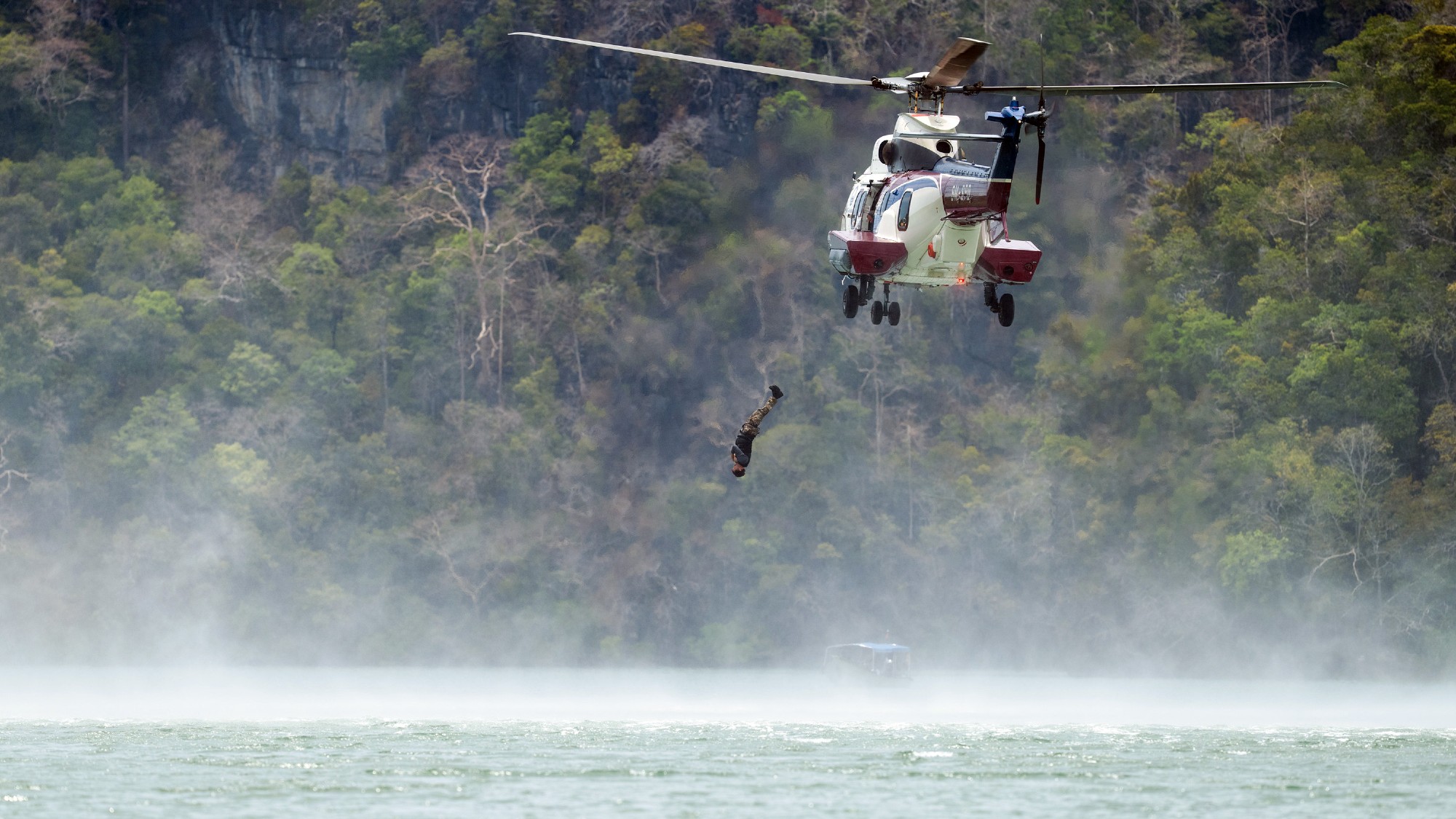 A recruit falls backwards out of a helicopter into the water. Trees are in the background and the water is light blue.