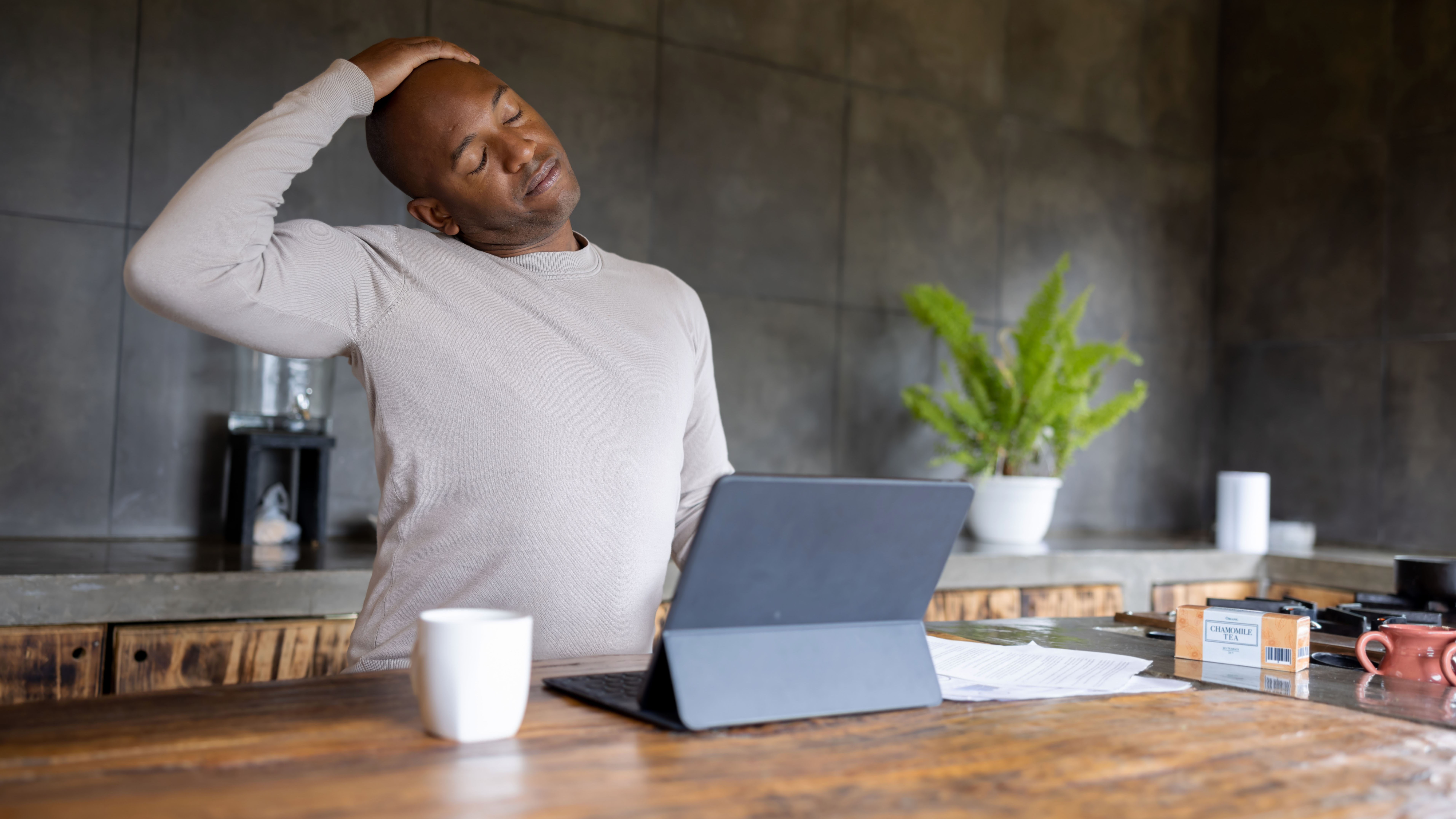 A man stands at a kitchen counter with a laptop open in front of him. He is stretching his neck by leaning his head towards his right shoulder. His right hand rests on top of his head, deepening the stretch. Behind him we see a blender and a potted plant.