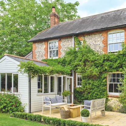 A brick and flint house with a large wisteria, patio with grey wooden benches and a fire pit beside the lawn.