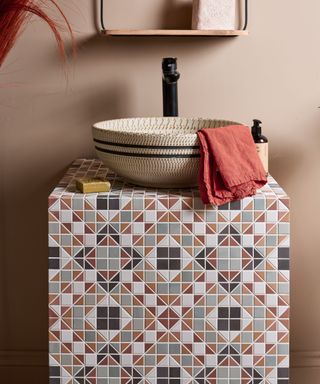 A countertop washbasin with a textured cream and black patterned design sits atop a bold geometric-tiled vanity unit. The tiles feature a mix of terracotta, sage green, charcoal, and cream tones, creating an intricate mosaic effect.