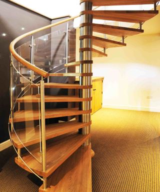 A timber, glass and steel spiral staircase from Complete Stair Systems. The staircase sits within a room with natural carpet and a mix of black and white walls.