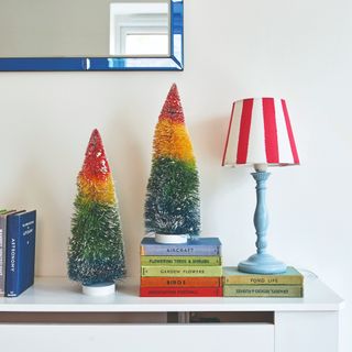 A radiator cover decorated with two miniature rainbow Christmas trees, books and a table lamp with a striped shade