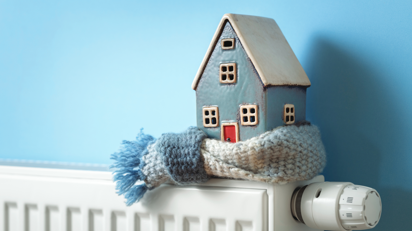 clay model of blue and white house with white and blue scarf knotted around base. house sat on top of white radiator with blue wall behind