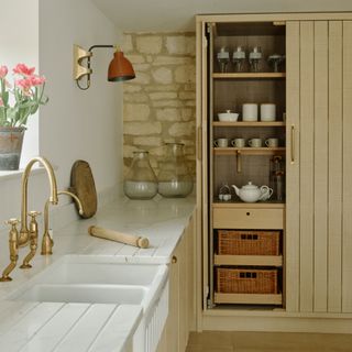 country kitchen ideas, country kitchen with exposed stone wall, pantry cabinet with open door, brass taps, butler sink, marble worktops, tulips on window sill, wall light