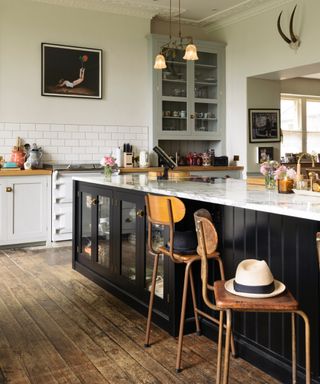 kitchen with black island with marble countertops, pale gray glazed cabinet, off white walls, white range, wooden countertops on white cabinet,s original wood flooring, vintage bar stools