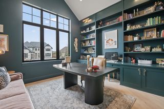 A home office with Sherwin-Williams' 'Cascades' paint on the walls with large windows, a beige sofa chair by a black desk and a wooden book shelf