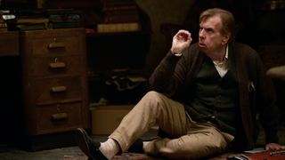 John Chapel (Timothy Spall) sits on the floor of his home, next to an old-looking mahogany desk and various piles of books and papers. He is looking up at someone or something unseen, and has the fingers of his right hand are semi-curled in concentration.