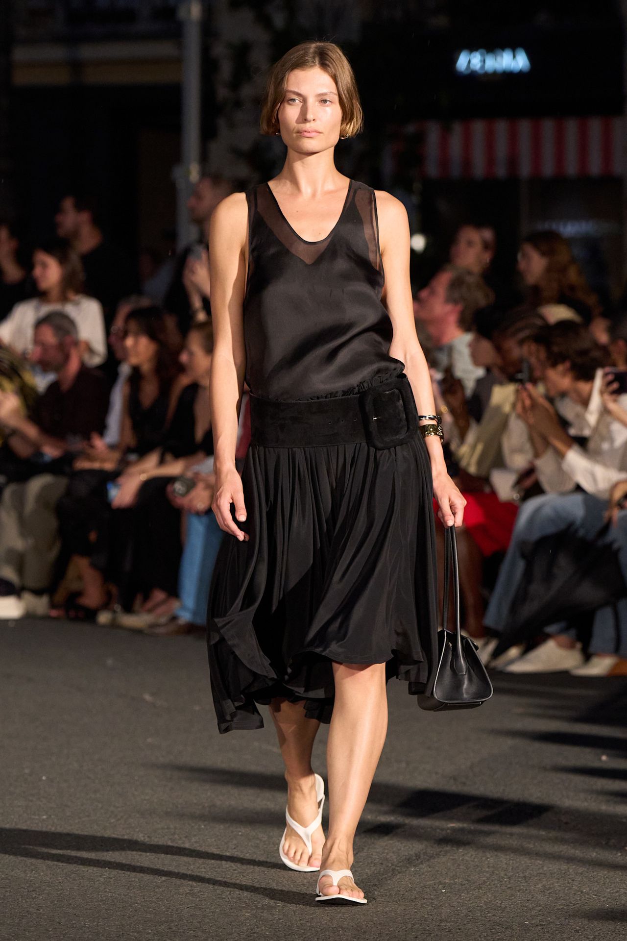 Layering staples for spring 2026 is shown in a photo from Ami Paris's runway show, with a model walking while wearing a black sheer organza tank layered on top of a black satin v-neck blouse tucked into a black midi skirt styled with an wide black belt, white flip-flops, and a black handbag