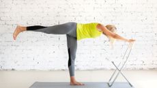 woman doing a balancing T shaped pose using a chair for support with a white wall background wearing yellow vest and grey leggings