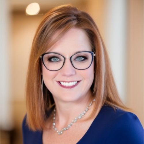 Laura Finson wearing glasses, blue top and silver necklace while smiling, against blurred out background.