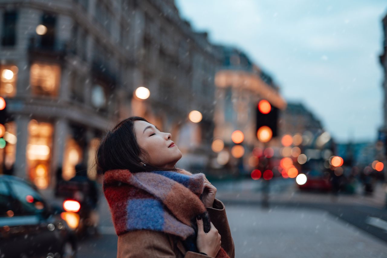 Woman in standing in a street at dusk with a city background.