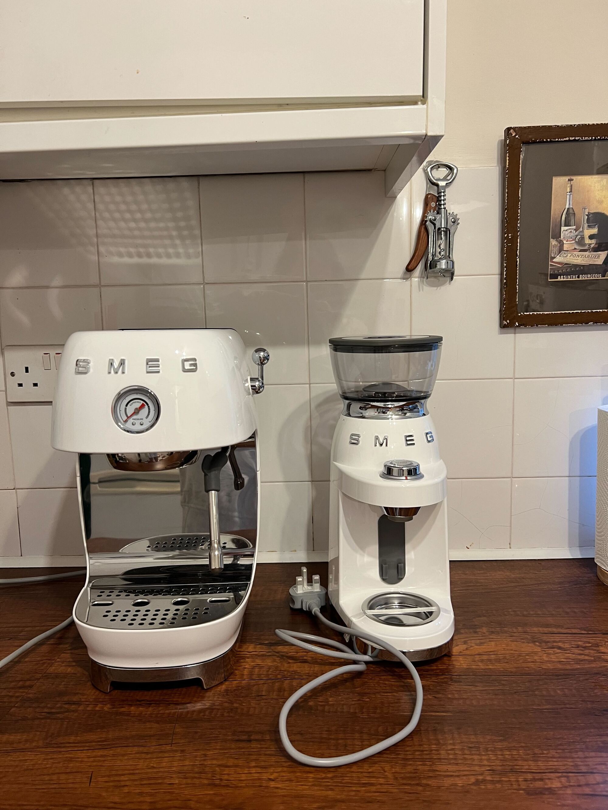 Image of a white Smeg cold brew machine and matching coffee grinder on a wooden countertop with white tile backsplash
