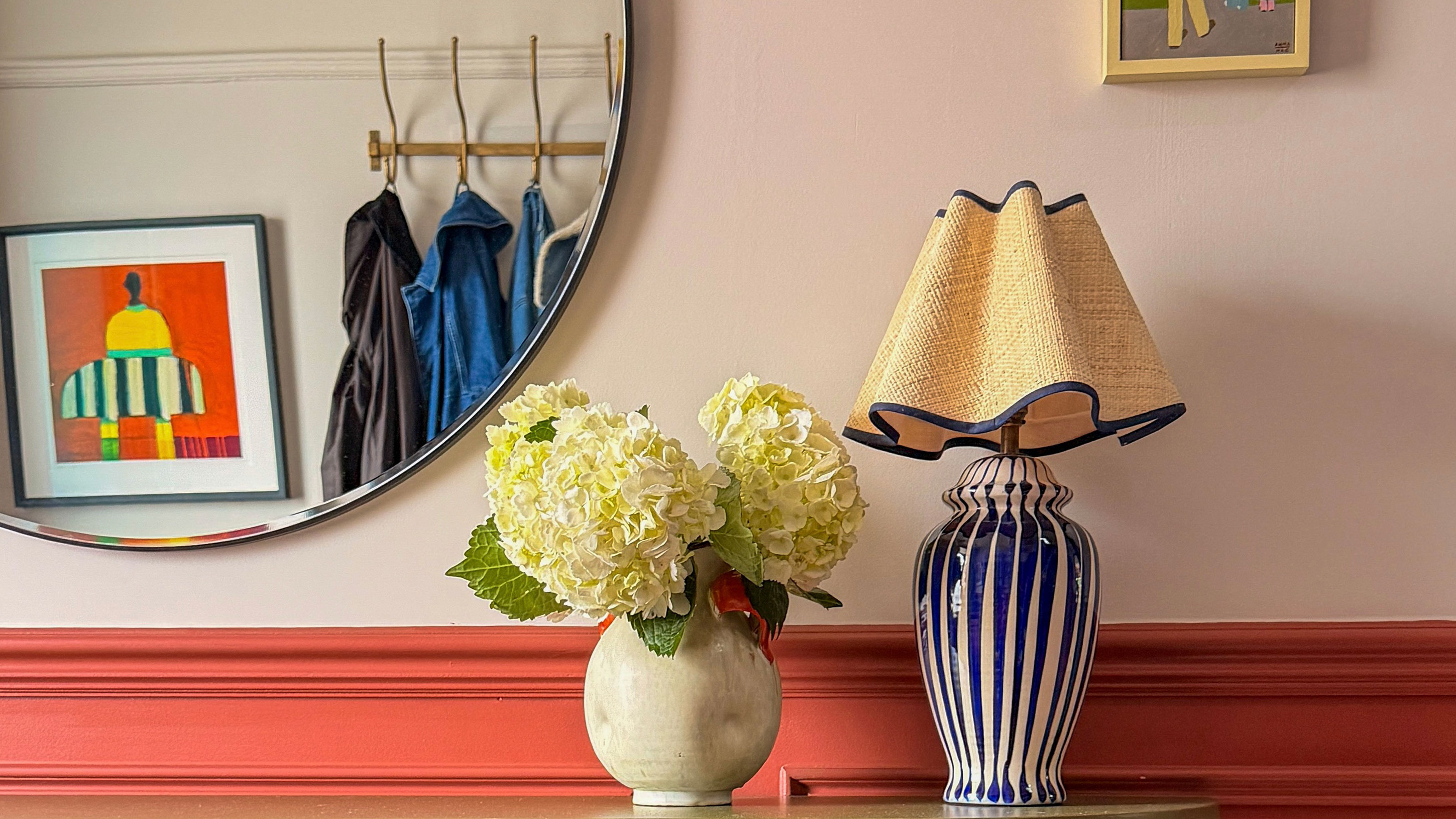 Hallway with lower half painted coral and top salmon, with vase of hydrangeas and blue and white striped lamp on shelf, beneath round mirror reflecting coat rack