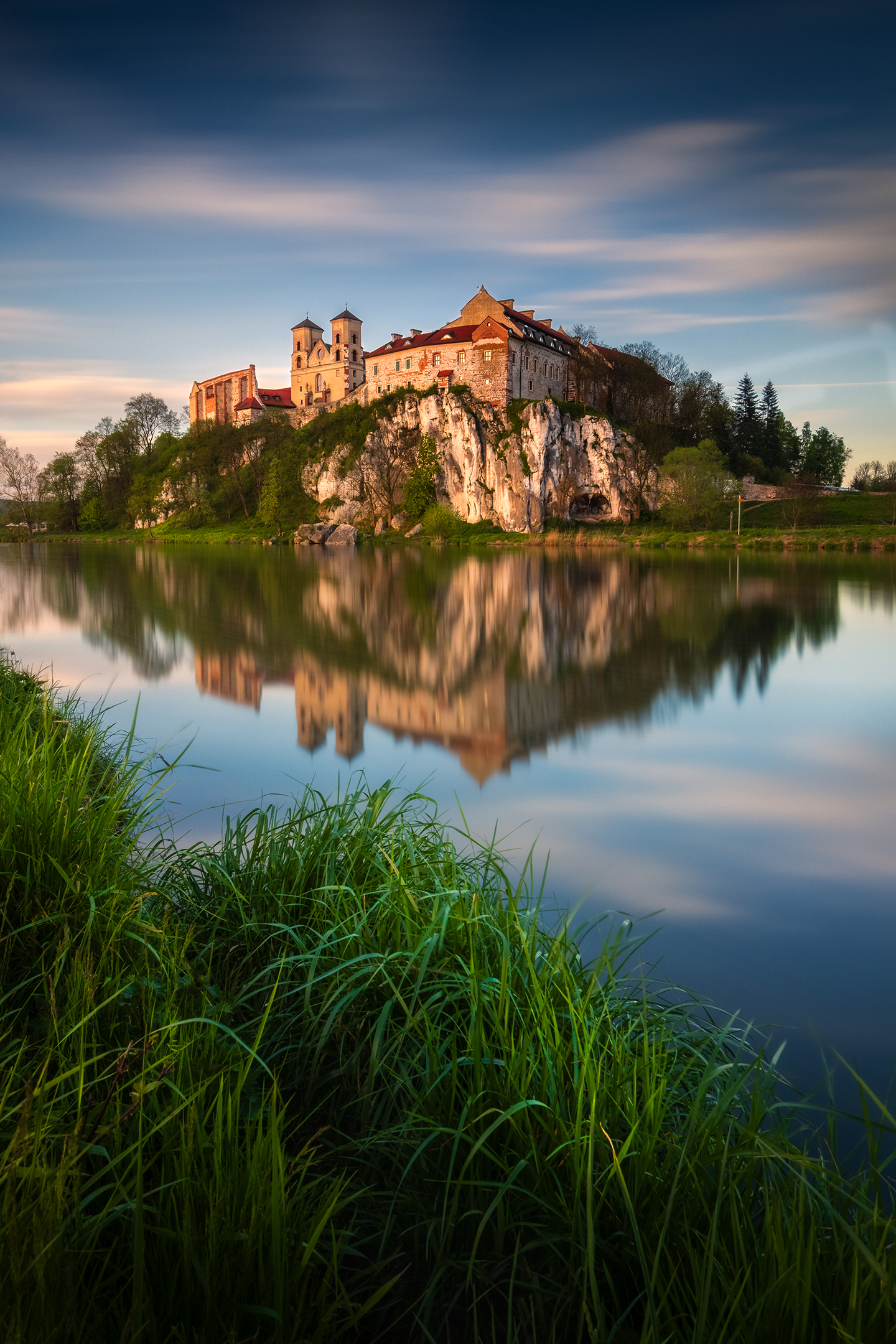 A historic castle perched on a rocky cliff, reflecting in calm waters, surrounded by lush greenery and a serene sky