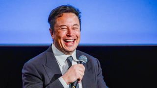 Elon Musk sitter på en scene med en mikrofon i hånden og ler.