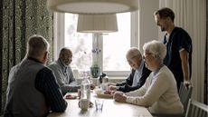 A group of assisted living residents smile and laugh over dinner while a caregiver stands nearby.