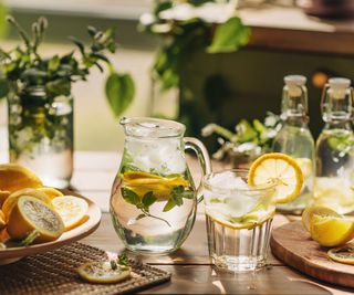 water pitcher in a table with lemon, mint, cups, and garnishes around it
